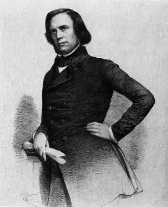 Noir et blanc, plan américain, en costume long croisé, chemise à col cassé blanche avec cravate noire dont le nœud n'est pas apparent. Regard ferme. Visage rectangulaire avec collier de barbe, cheveux bruns et longs, yeux foncés, nez en bec d'aigle.