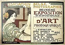 Affiche couleur annonçant la "Deuxième exposition internationale d'art photographique", illustrée par un femme aux cheveux noirs et vêtue d'une robe à fleur qui observe une photographie imprimée. Elle se trouve dans une salle où l'on observe de nombreuses photographies encadrées qui sont pendues au mur.
