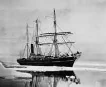 Photographie en noir et blanc du voilier Aurora en Antarctique. Des icebergs sont visibles.