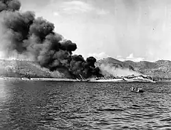 Photo noir et blanc d'un navire en flamme, avec une épaisse fumée noire et en train de couler