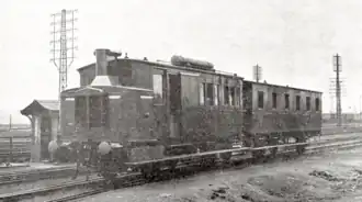 Photographie en noir et blanc de l'autorail de forme parallélépipèdique vue de trois quart face, côté gauche visible. Il est accompagné de sa remorque.