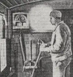 Photographie en noir et blanc de la cabine d'un autorail. Le conducteur est debout, porte un petit bonnet et tient dans sa main un levier. Sur la paroi de la cabine, un appareil enregistreur de vitesse est présent.