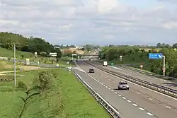 Une route 2x2 voies bordée des deux côtés par de l'herbe en premier plan et une aire d'autoroute en arrière-plan.