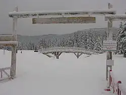 Clairière enneigée avec au premier plan un portail en bois et un insigne « Autrans : bienvenue à Gève, accès aux pistes », au second plan un ponton en bois et tout autour des conifères lourdement enneigés