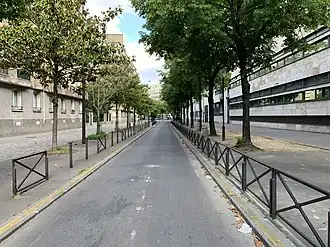 Photo d'une rue bordée d'immeubles de chaque côté, la chaussée est goudronnée et bordée de chaque côté par des barrières et des arbres.