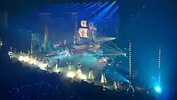 Photo d'une scène de concert où des chorégraphes sont alignés de front alors qu'un bateau de style ancien se trouve en arrière plan