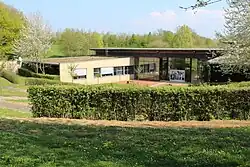 Bâtiment dont les couleurs font que ce dernier se fond dans le paysage dominé par des arbres verts et de l'herbe.
