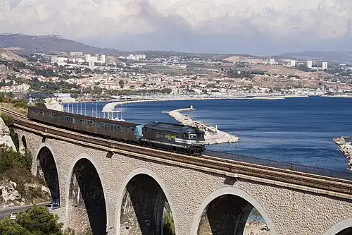  Un train passe sur un viaduc, à l'arrière plan un littoral urbanisé et la mer.