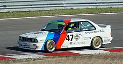 Photo vue de gauche d'une voiture blanche, avec deux rayures rouge et bleue, sur une piste de course