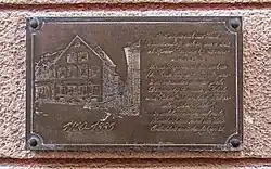 Sur la plaque figure une gravure de maison, à droite un texte descriptif.