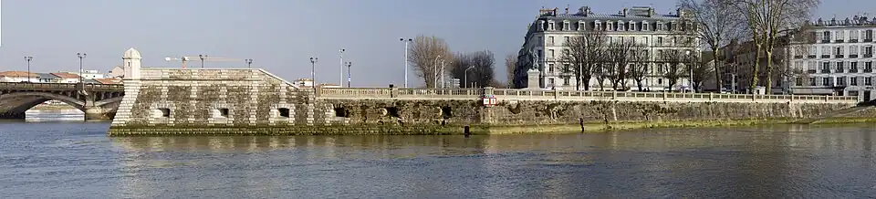 Vue d’un quai fortifié, orné d’une échauguette, en prolongement d’un pont sur un cours d’eau. À droite de la vue se dressent deux bâtiments imposants.
