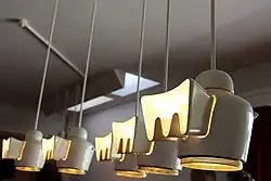 Luminaires de la salle à manger éclairant la Table ainsi que des tableaux au mur. Au fond on remarque l'éclairage indirect des puits de lumière remplissant la même fonction de jour.