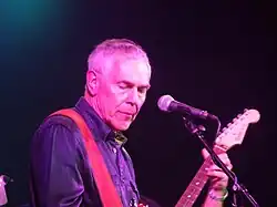 Photo en gros plan d'un homme blanc âgé portant une veste en jean. Il joue de la guitare devant un micro sur pied.