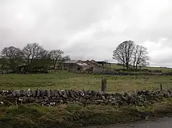 Ferme abandonnée, bâtiments gris, arbres nus, clôture de pierre, ciel nuageux. Paysage rural désolé, ambiance contemplative.