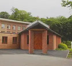 Photographie couleur montrant l'entrée d'un bâtiment bas en briques, surmonté d'une croix