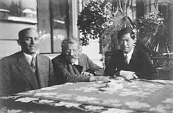 Photo en noir et blanc. On voit, de gauche à droite et autour d'une table : un homme qui se tient droit et sourit : Bernhard Schapiro, le directeur adjoint de l'Institut de sexologie ; un homme penché sur la table : Magnus Hirschfeld, directeur de l'Institut ; et un homme les bras croisés : Tao Li. Derrière eux, des plantes.