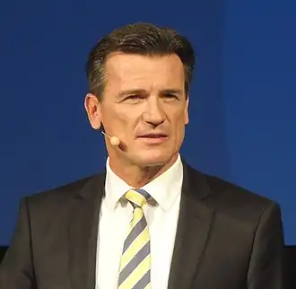 Photo de Bernhard Wolfgang en costume, vu de face devant un fond bleu.