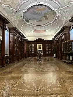 Photographie couleur d'une vaste salle comprenant de nombreuses vitrines en bois et un plafond décoré de stuc blanc et de fresques.