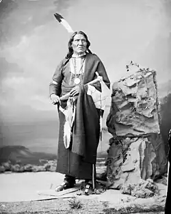 Photographie en noir et blanc d'un Amérindien en habit traditionnel coiffé d'une plume et tenant un tomahawk.