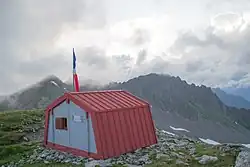 Petit refuge de montagne en tôle rouge avec des montagnes en arrière-plan et un ciel nuageux.