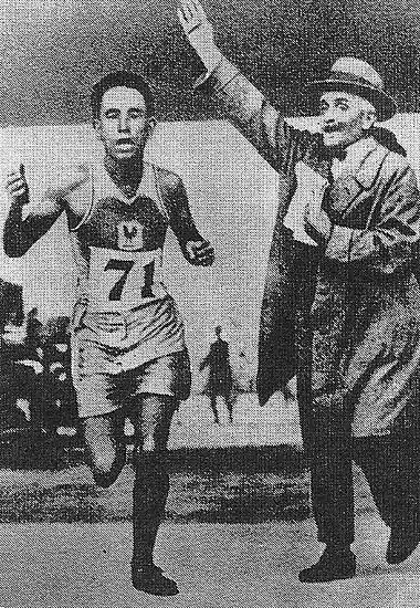 El Ouafi, avec le dossard 71, franchit la ligne d'arrivée devant un juge portant un manteau et coiffé d'un chapeau avec le bas droit levé et des notes dans la main gauche.