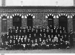 Photographie noir et blanc d'étudiants en quatre rangs avec leurs professeurs, devant un bâtiment en brique.