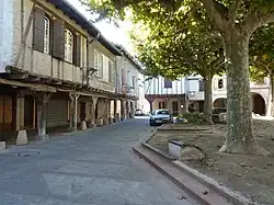 photo couleur de maisons à colombage avec avancée sur couvert au rez-de-chaussée. La place est ombragée par des platanes.