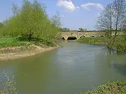 Au premier plan, une rivière aux rives basses avec quelques arbres. En arrière plan un pont de pierre dont on voit 3 arches