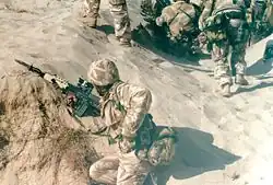 photographie en couleur montrant un soldat accroupi dans une tranchée de sable ; il porte un uniforme camouflé beige et un fusil d’assaut avec un baïonnette fixée au bout du canon