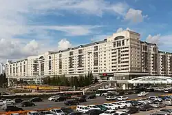 Vue d'un long bâtiment blanc aux colonnes noires. Au premier plan un parking avec des voitures et une route passante.