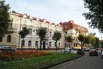 Bâtiment néoclassique de couleur crème, jouxté d'autres bâtiments au bord d'un trottoir arboré.