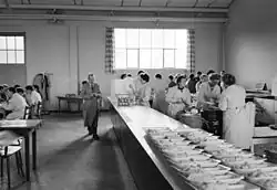 Photo en noir et blanc de l’extrémité d’une grande salle. À droite, sur un comptoir, une vingtaine de plats partiellement servis attendent d’être complétés par des femmes en tablier blanc. Au centre, un homme moustachu, en tablier de travail, s’avance parallèlement au comptoir et semble rouler une cigarette. À gauche, quelques hommes attablés mangent et discutent.