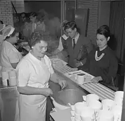 Photographie en noir et blanc. Des femmes en uniforme blanc servent les portions de nourriture sur les plateaux qu’étudiants et étudiantes font glisser devant elles sur un comptoir rainuré. Les étudiants sont en costume-cravate, les étudiantes sont élégamment vêtues.