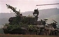 photographie montrant un Gepard de face et couvert de végétation pour le camoufler, suivi de plusieurs véhicules blindés à roues