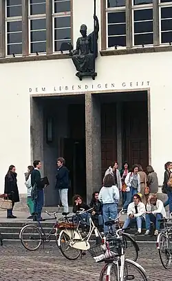 L'entrée d'un bâtiment blanc devant laquelle discutent des étudiants. Au premier plan, des vélos.