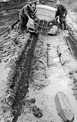 Photo en noir et blanc. Plusieurs soldats poussent une voiture embourbée. Des traces de roues montrent la boue sous la neige.