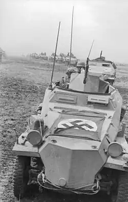Photo en noir et blanc d'une colonne de véhicules blindés. Celui au premier plan porte un drapeau nazi sur le capot.