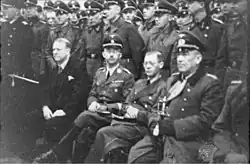 Quatre hommes sont assis devant des rangées de soldats au garde-à-vous. L'homme le plus à gauche est en costume, les autres en uniforme.