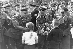 Photographie montrant deux enfants, une fille à gauche et un garçon à droite remettant un bouquet de fleurs à Heinrich Himmler au centre de la photographie ; le personnage de gauche est Wilhelm Koppe, celui de droite Fritz Bracht.