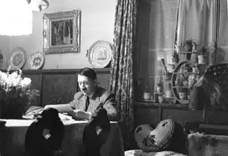 Photo noir et blanc montrant Adolf Hitler, assis à une table, dans une pièce de sa résidence secondaire du Berghof (à Berchstesgaden). L'homme, en costume clair, de face, est en train de lire des documents. L'arrière-plan est divisé par un rideau aux motifs fleuris : à droite, une fenêtre fermée au bord chargé de pots de plantes et, à gauche, un mur blanc supportant un tableau et un meuble en bois d'une couleur foncée sur lequel sont posées, sur la tranche, trois assiettes.