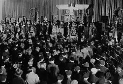 Photographie en noir et blanc d'une foule écoutant un discours d'un orateur placé sous des symboles nazis.
