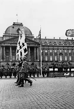 Photographie en noir et blanc montrant des soldats alignés au premier plan, vus de profil avec le palais royal à l'arrière-plan, un drapeau nazi flottant dans les airs en haut à droite du cadre.