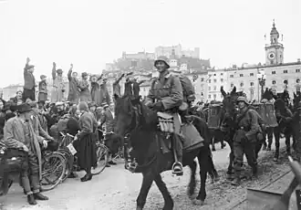 Des soldats passent à cheval devant une foule réjouie qui leur adresse des saluts nazis, certaines personnes tiennent des fanions à croix gammée. En arrière-plan, la citadelle de Salzbourg et des bâtiments historiques.