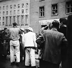 Photo en noir et blanc d'un groupe d'ouvriers du bâtiment, de dos, tournés vers un rang d'hommes en uniforme à l'arrière-plan