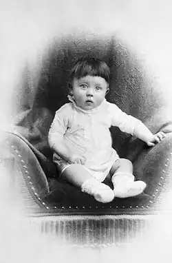 Photo en noir et blanc d'Adolf Hitler prise peu après sa naissance. Sur un fond blanc, dans un médaillon au fond sombre, au centre de la photo, un bébé, en layette blanche, est assis sur un siège en velours foncé. Les yeux grands ouverts, il fixe l'appareil photo avec curiosité.