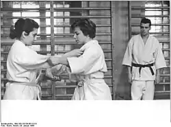Photo en noir et blanc de deux femmes en judogi et ceinture claire s'affrontant sous le regard d'un homme en judogi et ceinture noire.