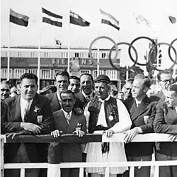 Un homme âgé en fustanelle entouré de nombreux autres hommes en costume.