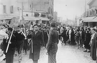 Photographie en noir et blanc d'hommes en civil portant des pelles, marchant dans une rue sous la surveillance d'un soldat.