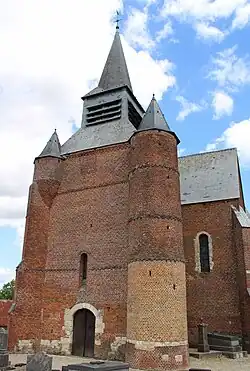 La façade de l'église. À droite une haute tour percée de meurtrières ; à gauche une échauguette en brique assise sur un imposant contrefort également en briques.
