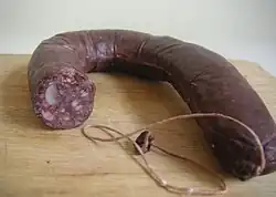 Sur une planche en bois, un morceau recourbé de boudin noir ; une extrémité est fermée par une ficelle, l'autre, tranchée net, montre les morceaux de graisses et la pâte de viande qu'enferme la peau de la charcuterie.